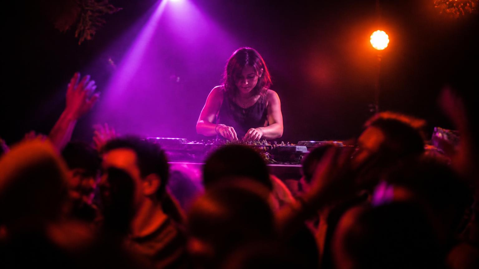 DJ tocando em uma pista escura iluminada por luzes roxas e âmbar